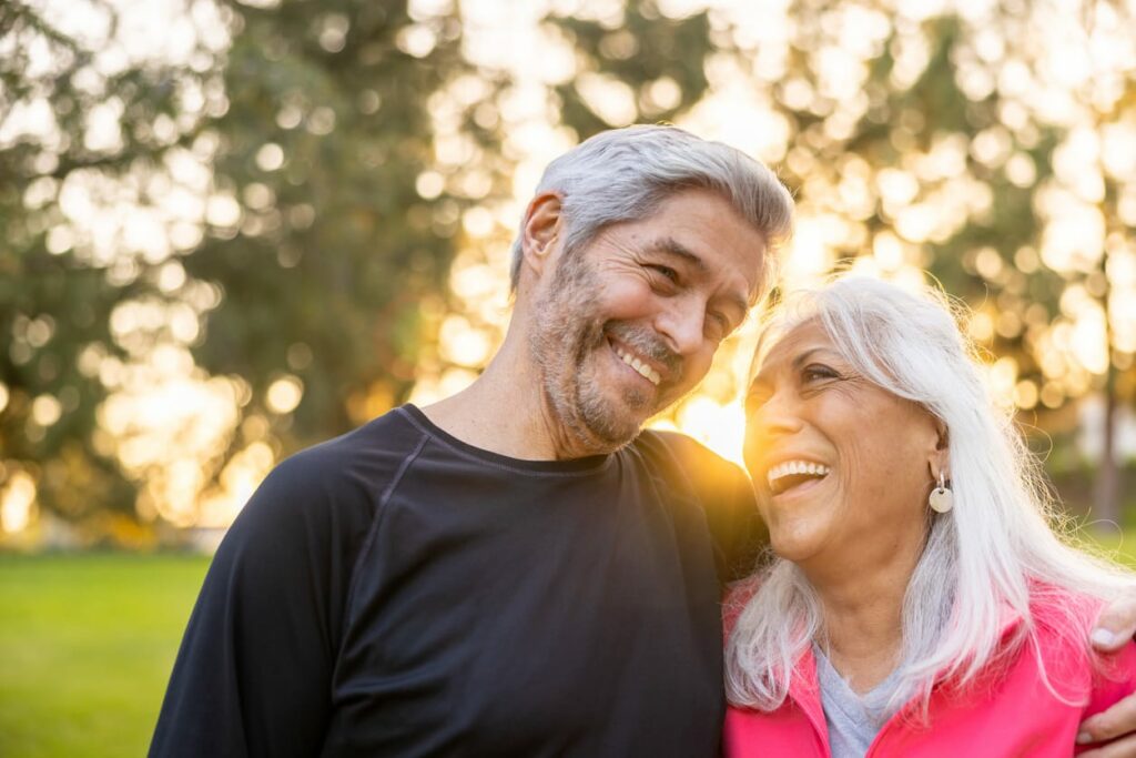 Two senior adults embrace and laugh while standing outdoors during sunset.