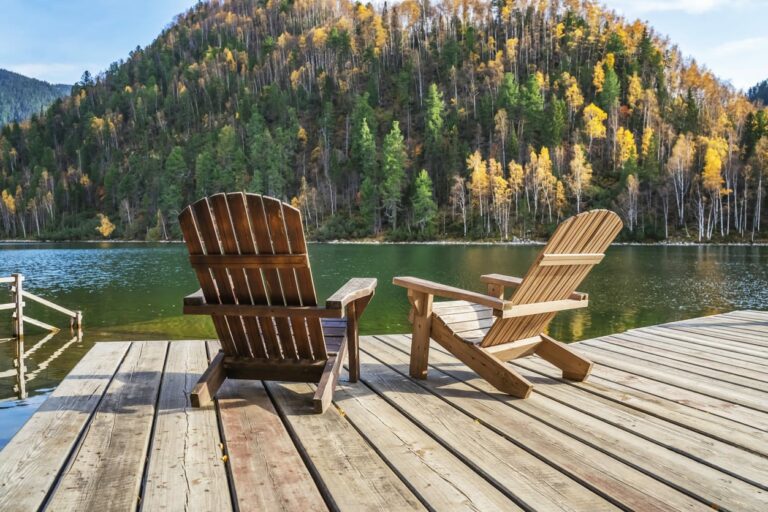 Two adirondack chairs sitting on a dock at the edge of a lake overlooking mountains with pine trees.